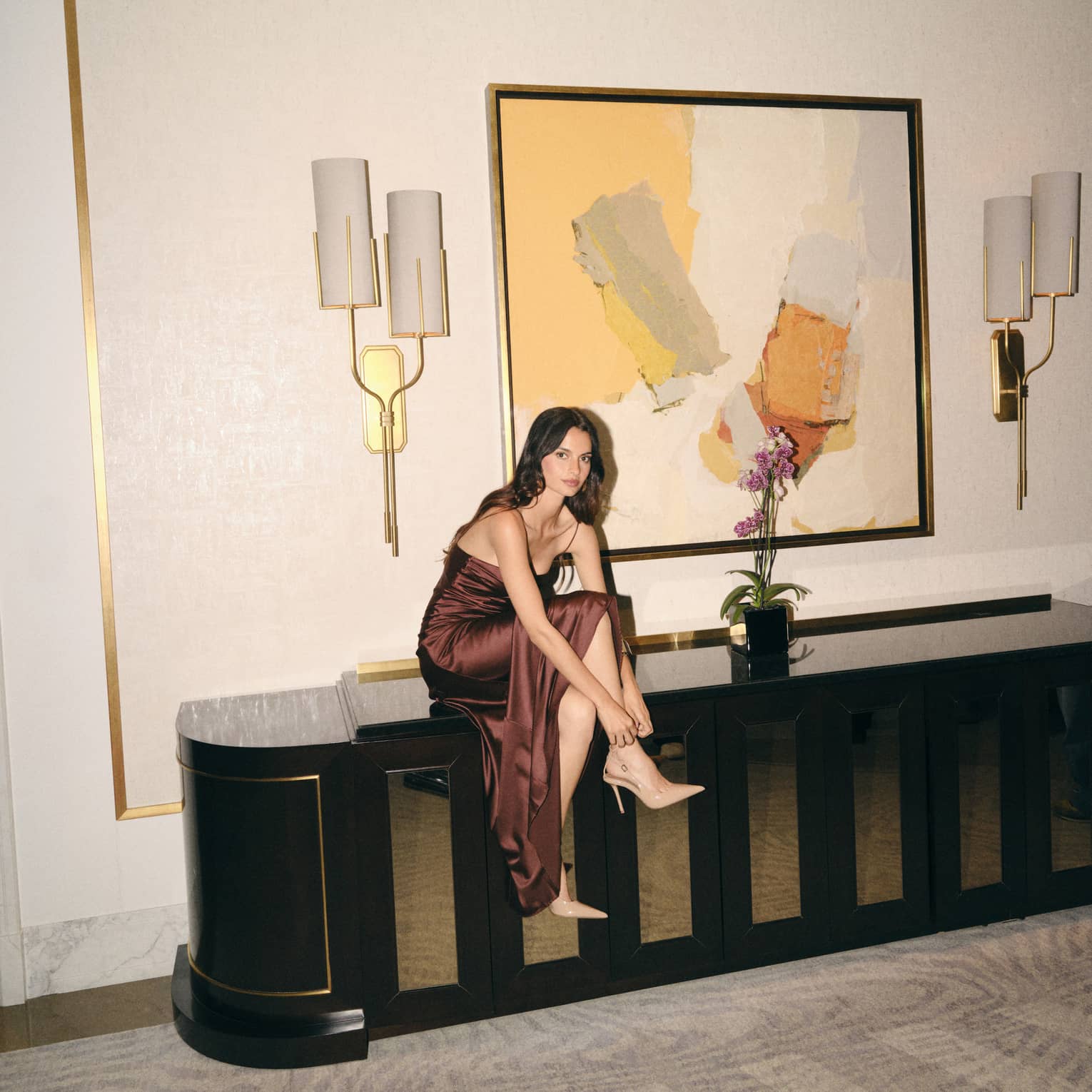Woman in formal dress sits on bench, fixing high-heel shoe
