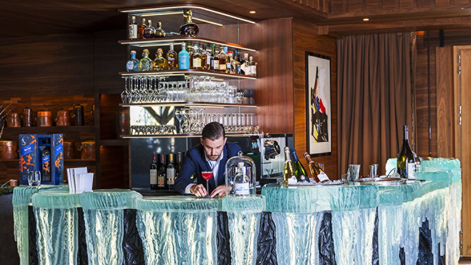 In a wood-panelled room, a bartender in a suit carefully adjusts a cocktail on a glass bar that looks like icicles.