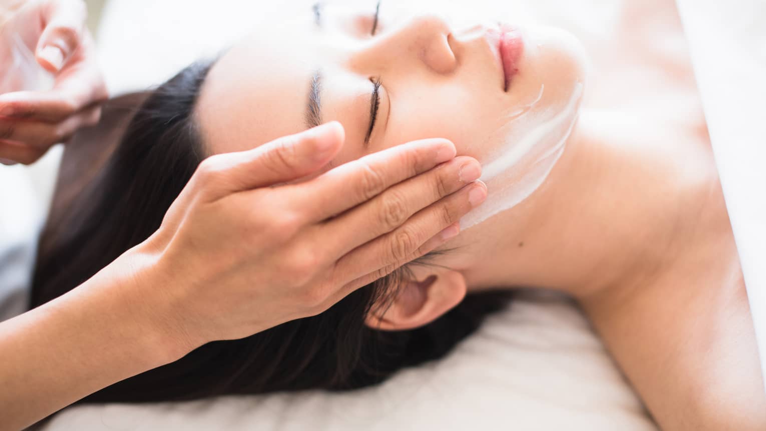 Close-up of spa attendant's hands rubbing white cream on woman's face as she lies on massage table