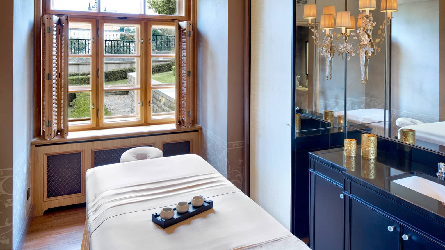 Tray with three small bowls on massage table with white blanket in front of window with open shutters