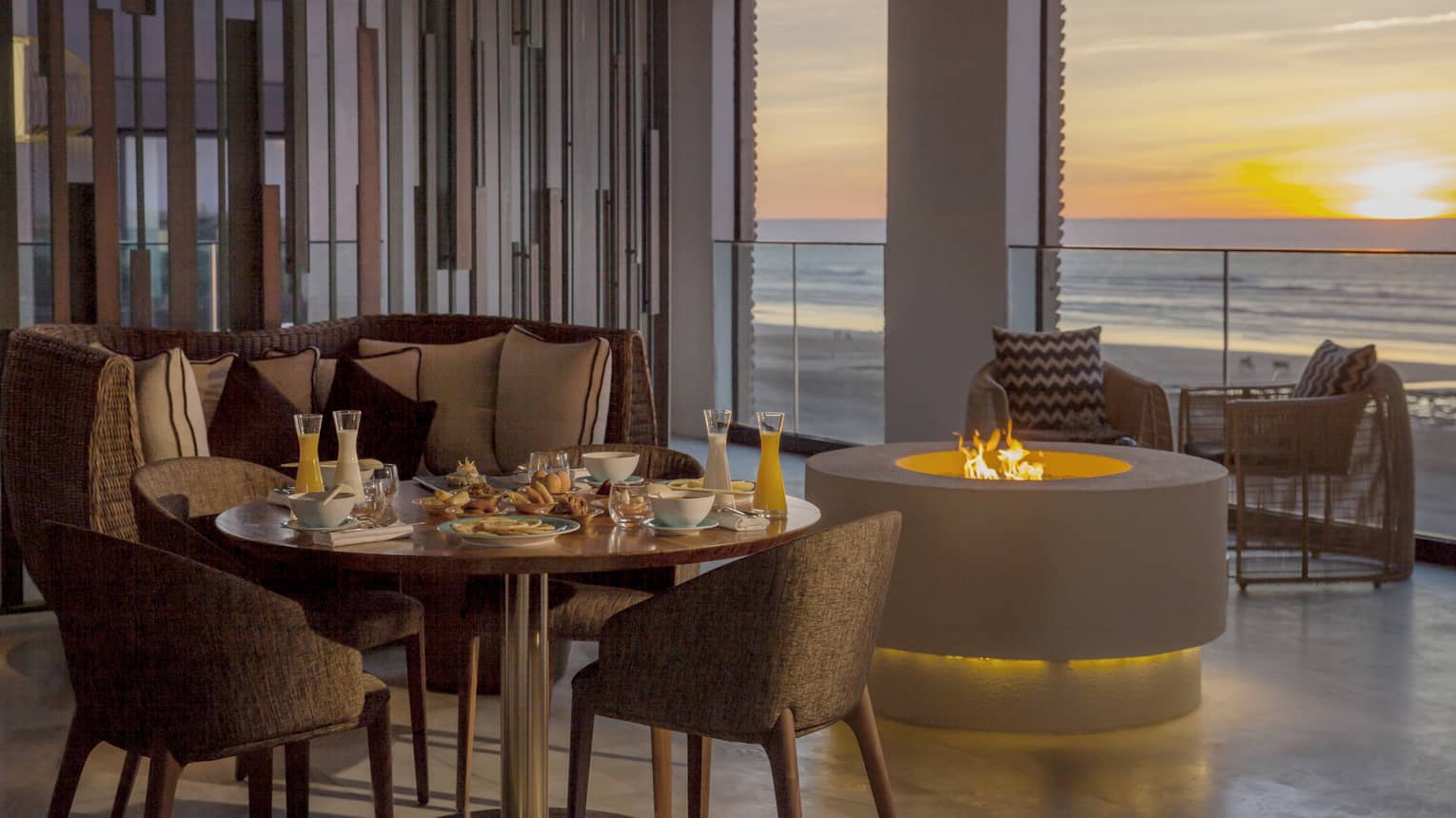 Restaurant dining table next to small modern firepit by glass balcony overlooking sea, sunset