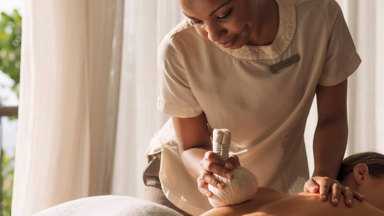 Woman holds stuffed round cloth over woman's bare shoulders as she lies on massage table