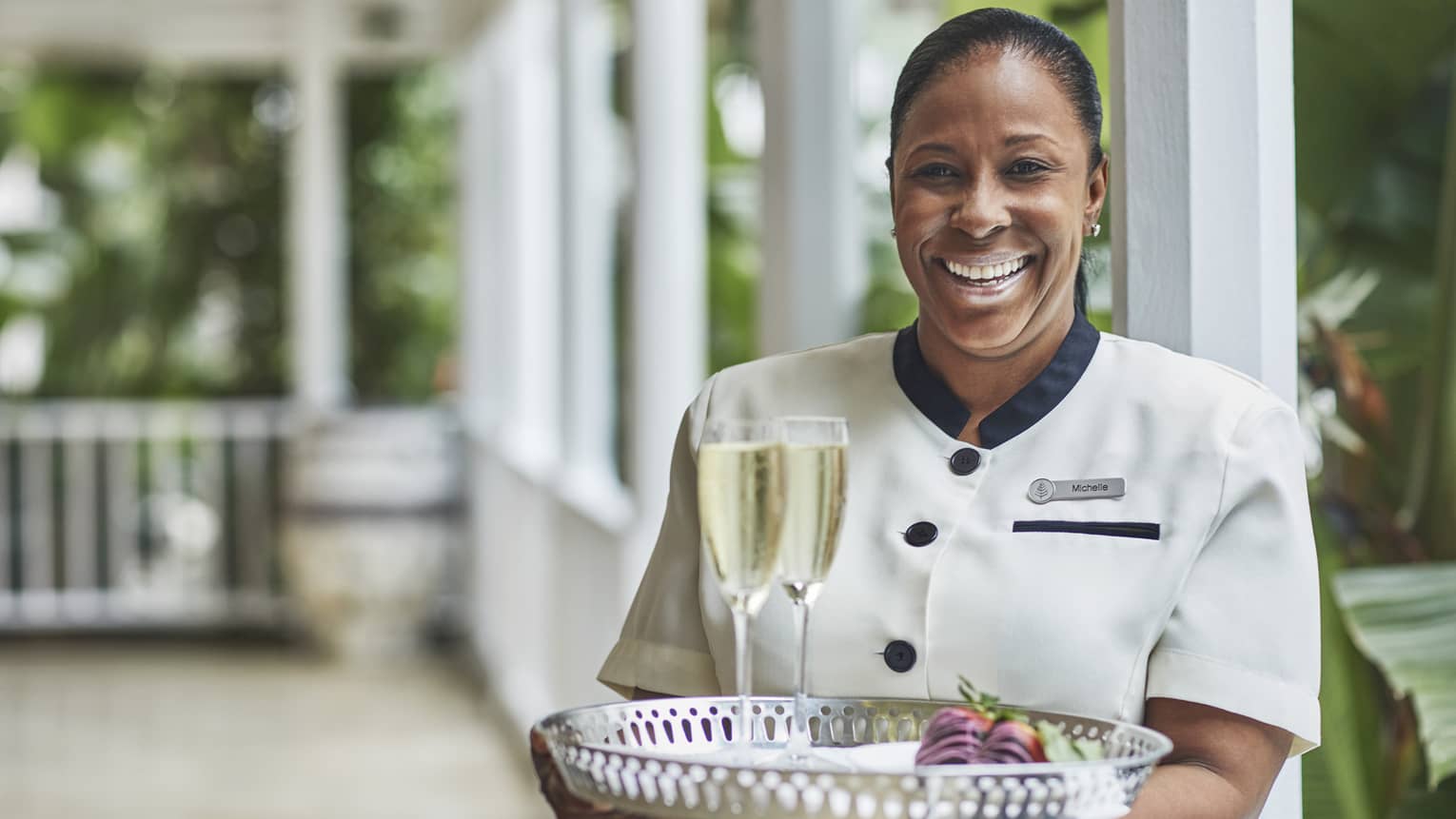 Smiling hotel staff holds tray with two glasses of Champagne on veranda
