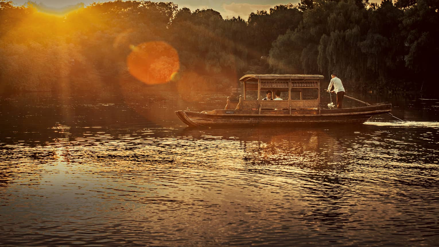 Man steers Chinese wooden rowboat on West Lake, into light of orange sunset
