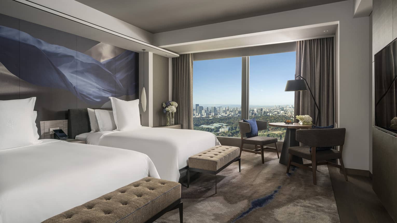 Guest room with two beds, tufted benches, seating area and floor-to-ceiling windows offering city views