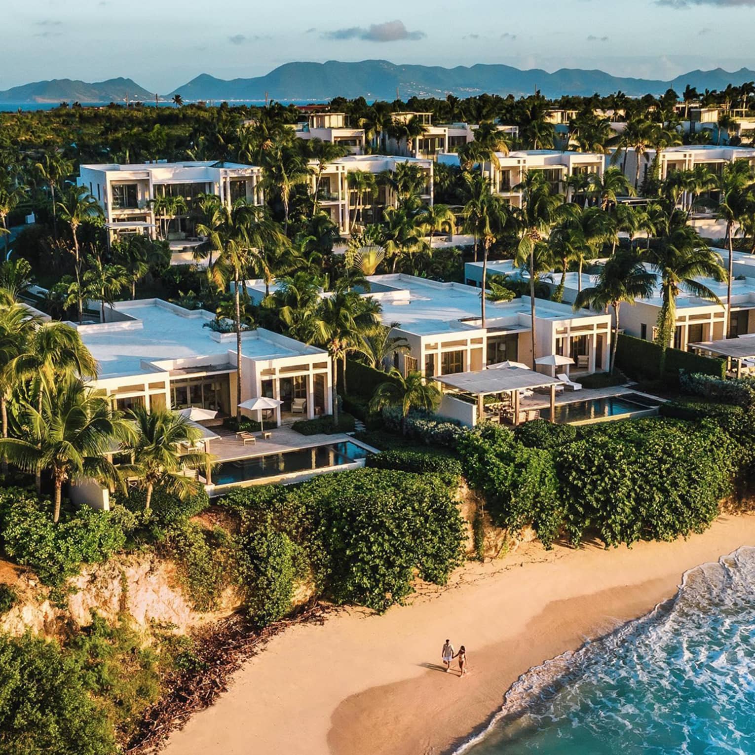 Aerial view of luxury resort beachfront villas surrounded by palm trees, with two people walking on the sandy beach below and mountains visible in the distance