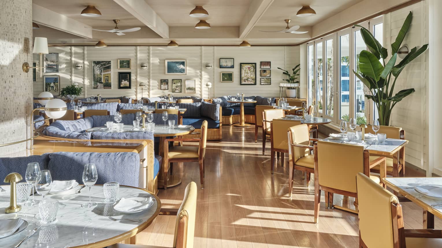 A sunlit dining outlet with large windows, wooden chairs and tables with blue accents and paintings on the wall.
