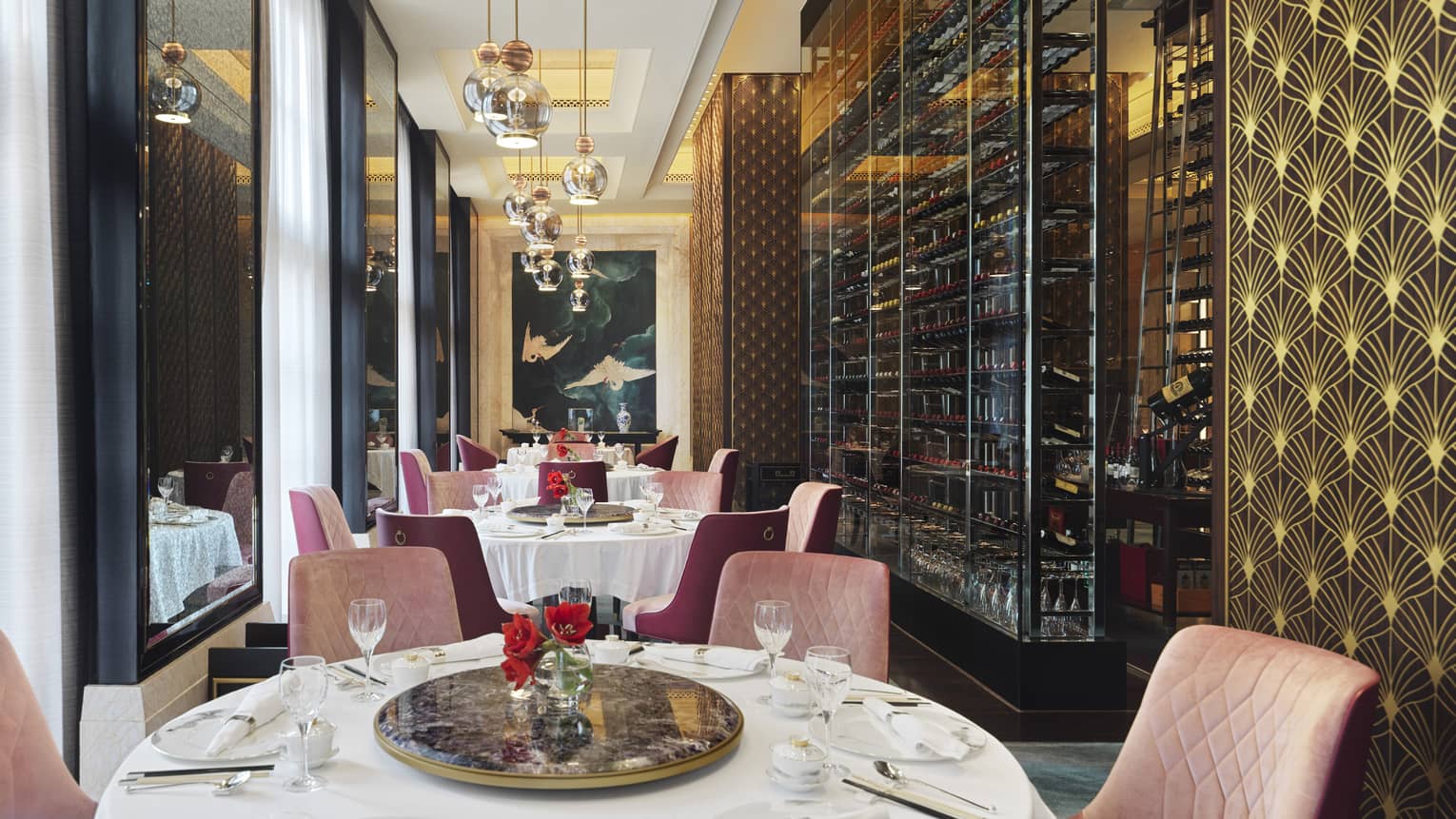 Opulent dining room with gold and pink accents, iron wine rack covering one wall, mirrors opposite, and a painting of cranes.