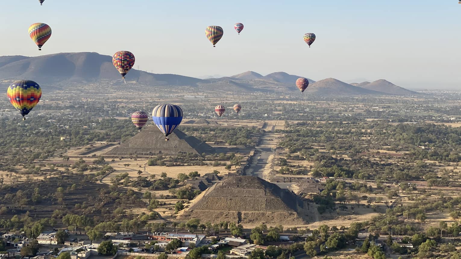 Hot-air balloons floating over old temples in a dessert covered in shrubs.