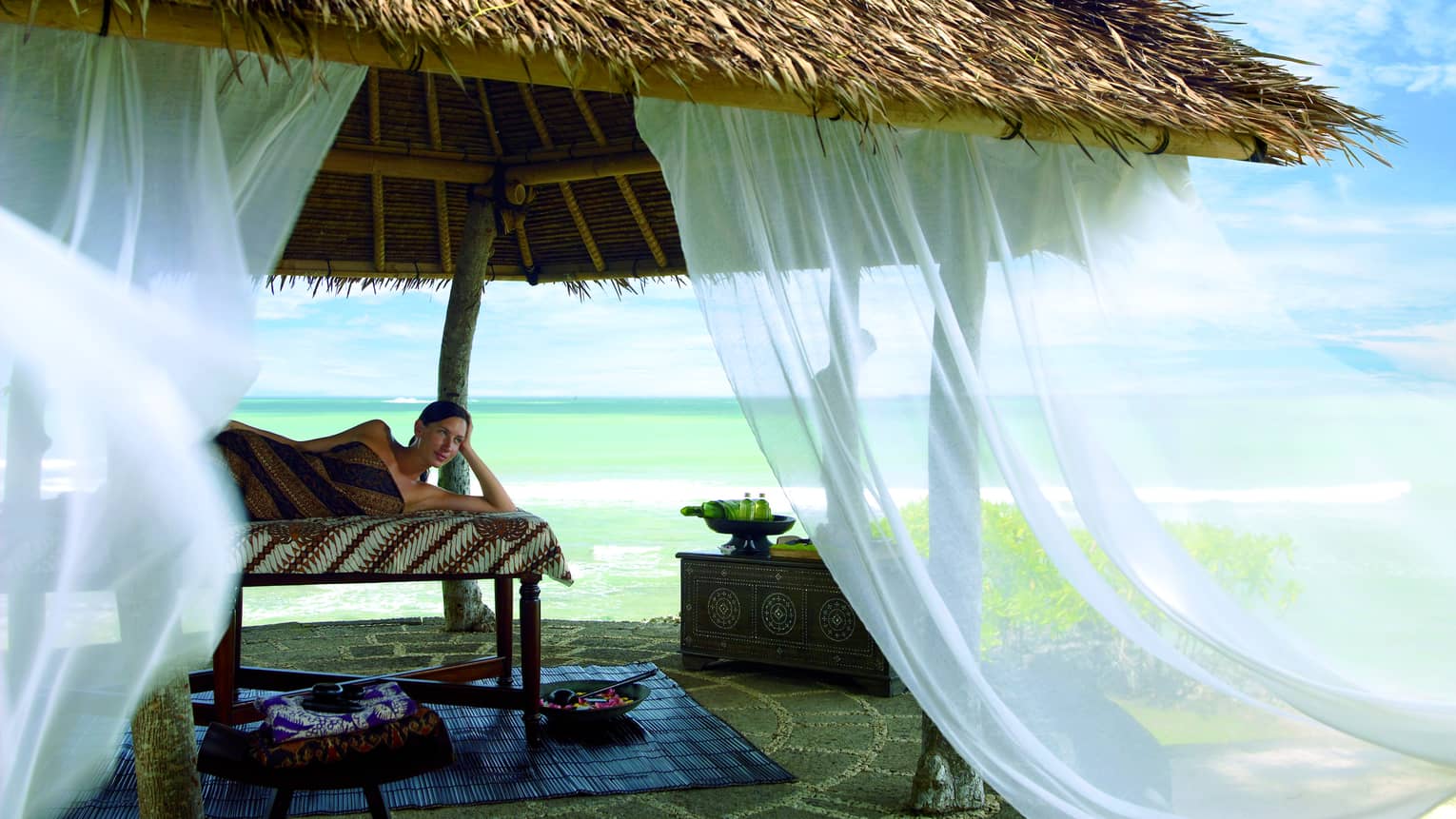 White curtains flow in the wind off thatched-roof gazebo as woman lies on massage table, gazes out at beach