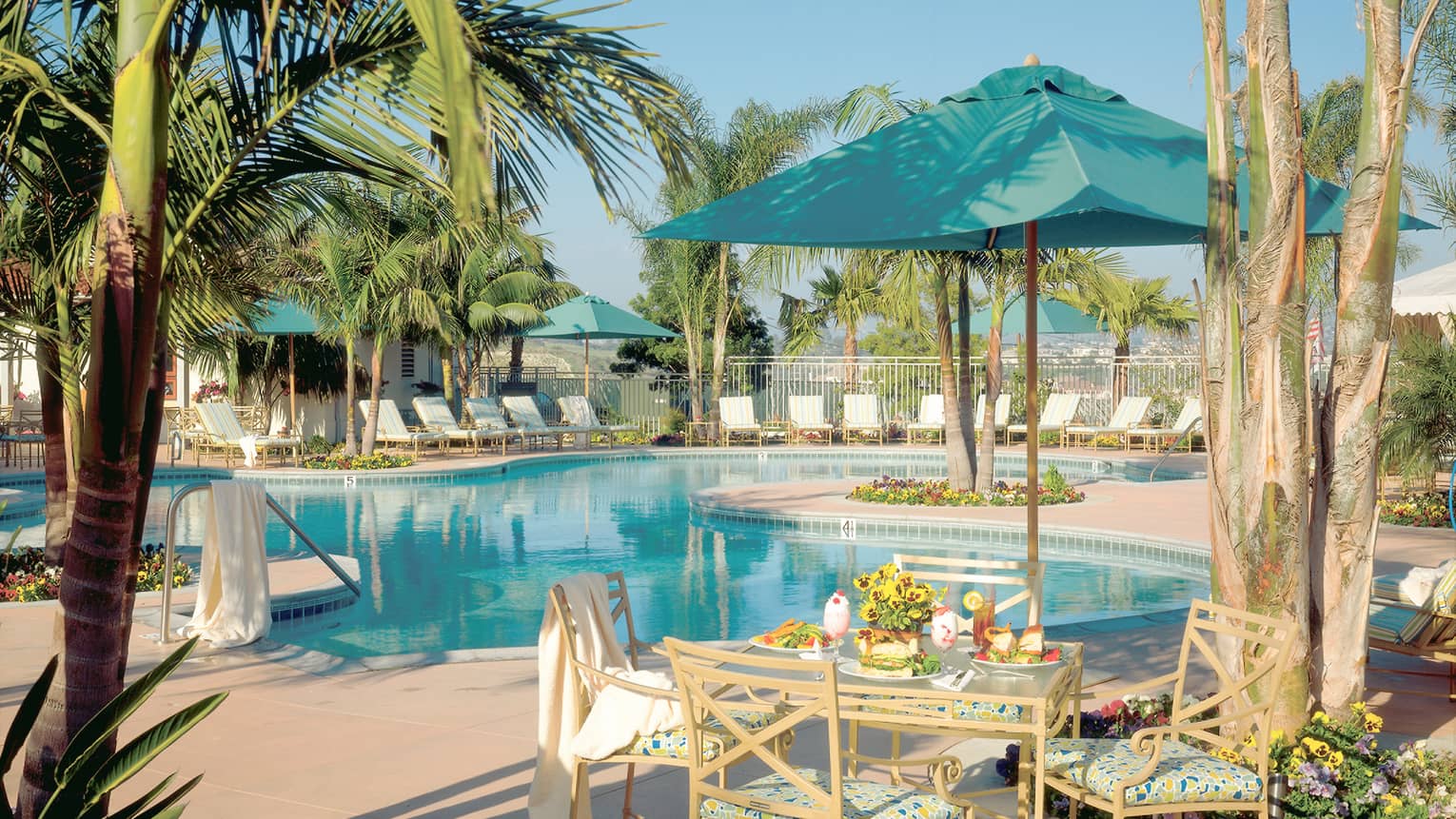 Outdoor pool and pool terrace, white lounge chairs, a square table and four chairs, turquoise umbrella, palms