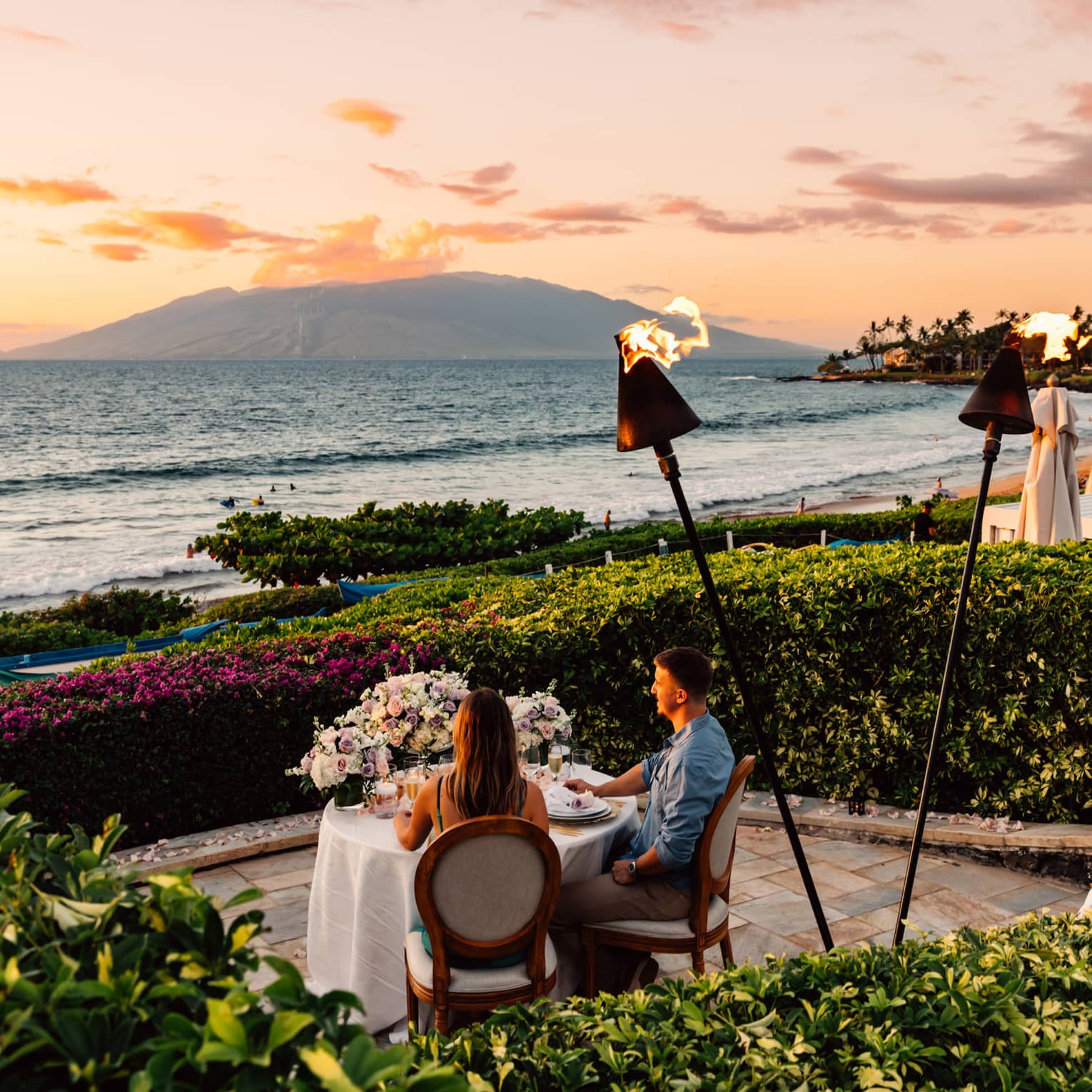 Couple dining at a table by the ocean at sunset with tiki torches and floral centerpiece