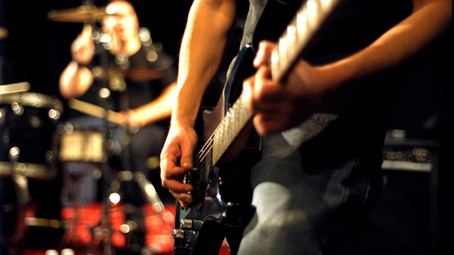Close-up of musician's arms and hands playing an electric guitar with drumset in the background.