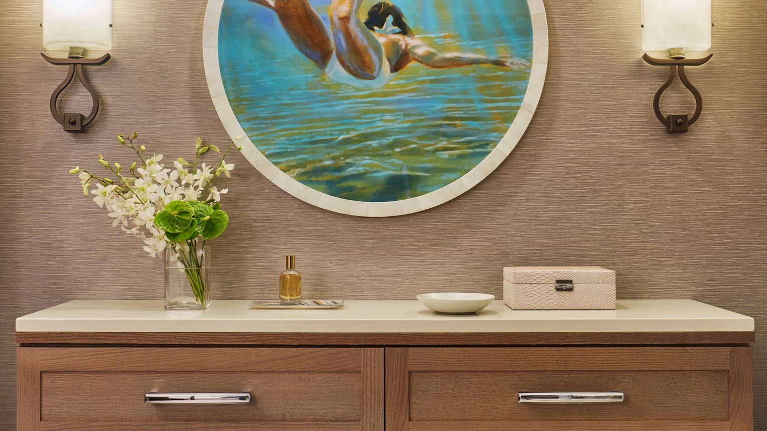 Wall-mounted console with flowers, perfume and decor beneath round artwork of a swimmer and two wall sconces