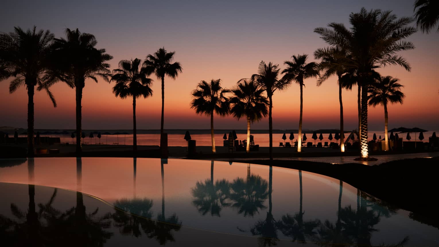 A palm tree?lined pool with the beach in the background all lit under a dark orange and blue sky as the sun sets