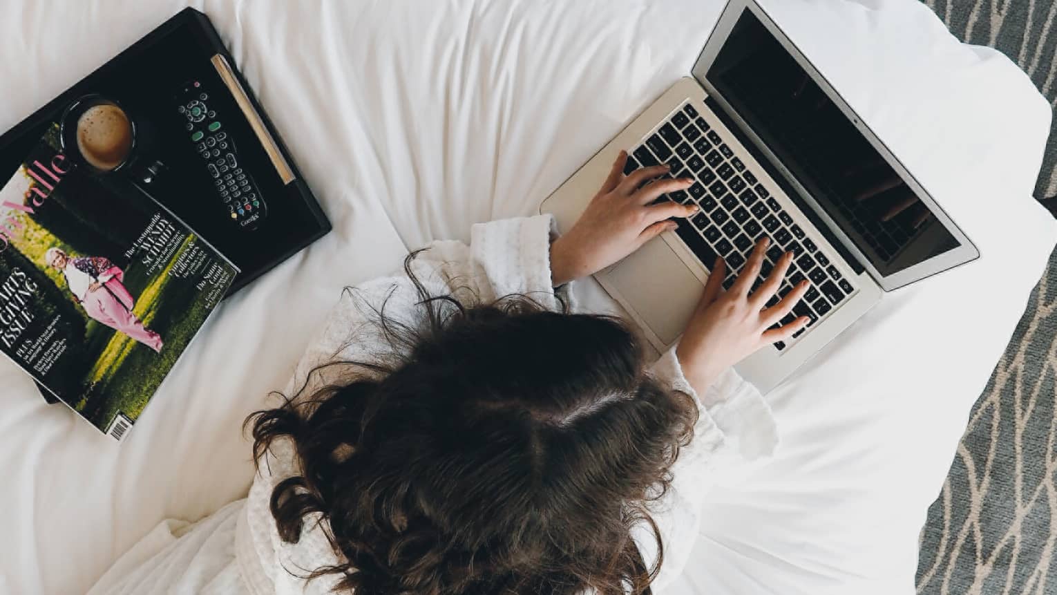 Aerial view of woman in bathrobe working on laptop on bed, stack of magazines
