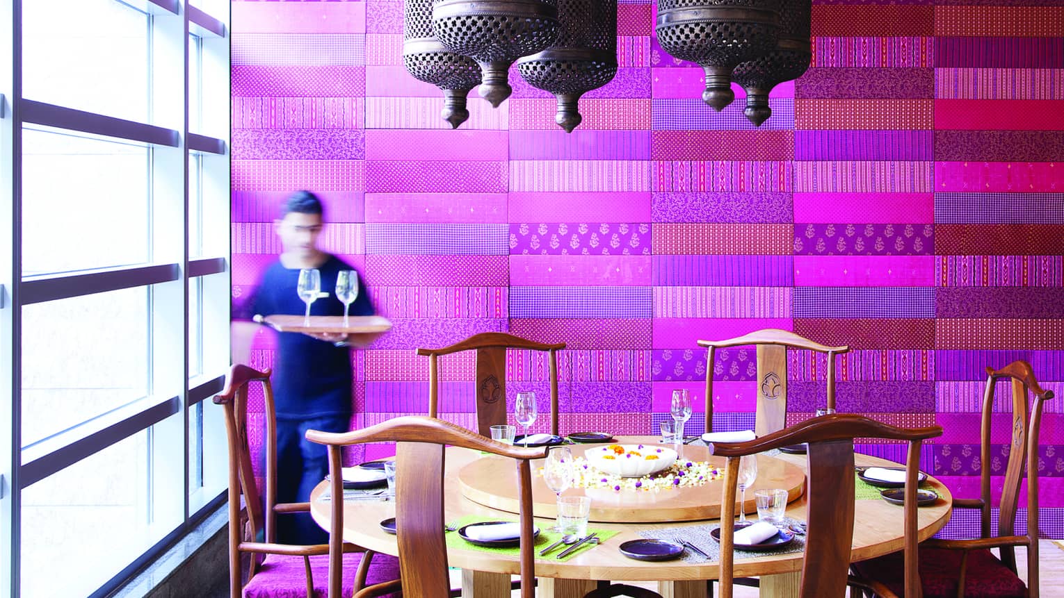 Waiter holds tray with wine glasses by large round dining table in front of purple textile wall