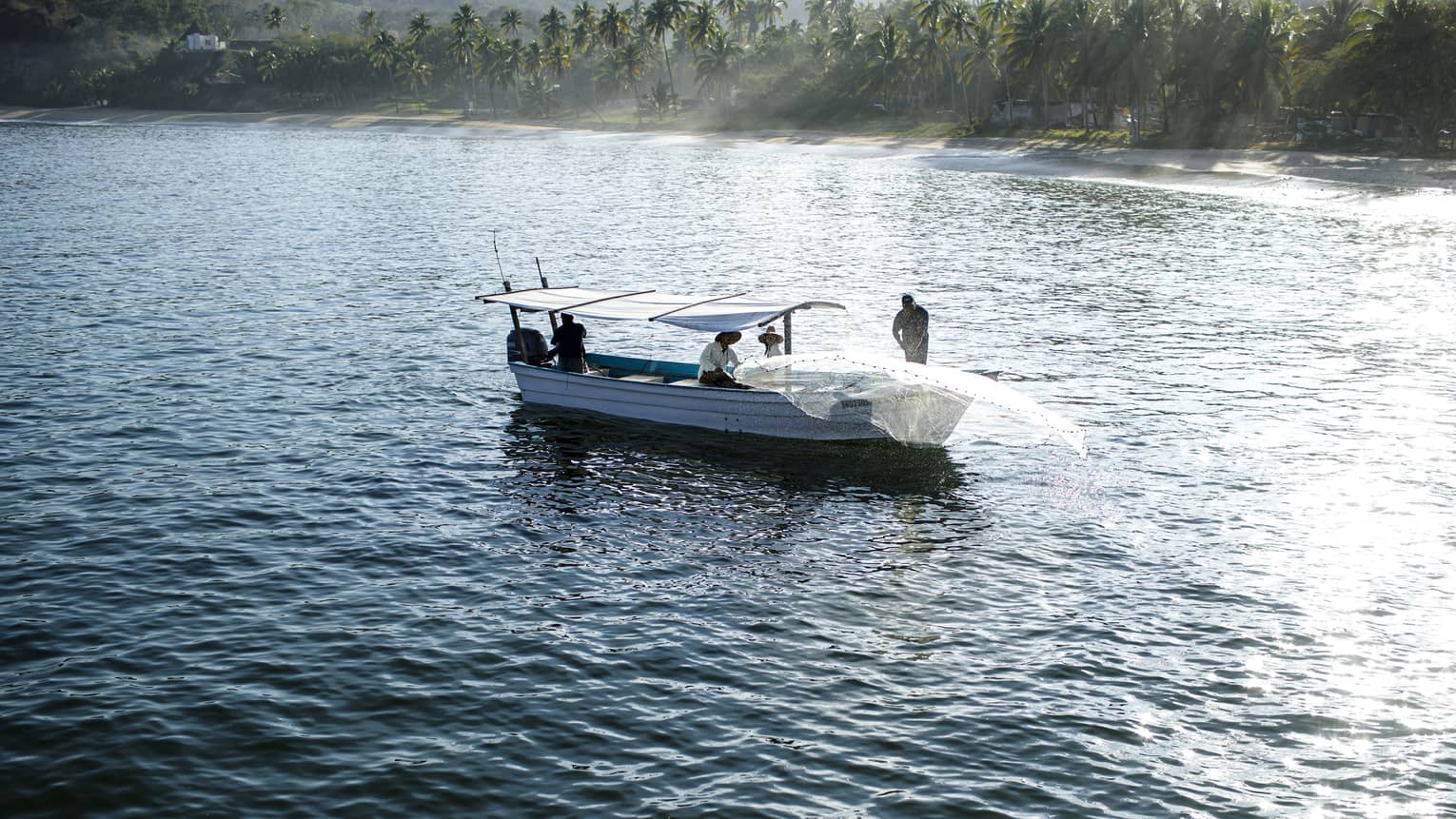 Long-view of a net being cast from the bow of a boat, three others under a canopy, daybreak streaming over palm trees beyond.
