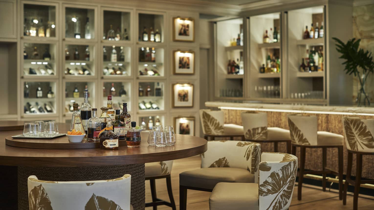 A round wood table set for a rum tasting in an elegant bar, backlit shelves around the room filled with bottles and glasses.