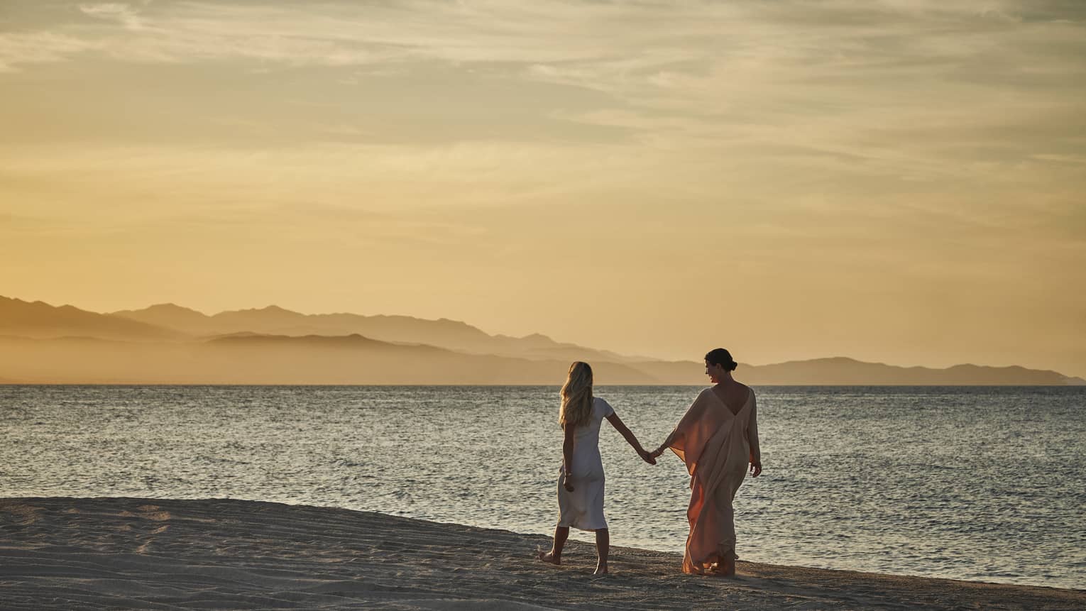 Two women holding hands and walking along a beach while the sun is setting.