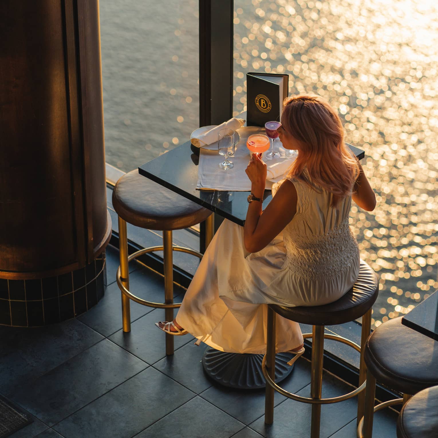 A person wearing a dress and sipping a drink while sitting at a dining table by a window
