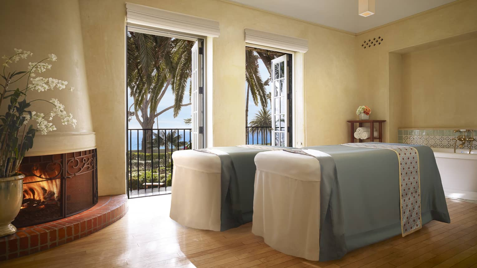 Two couples massage beds with blue sheets by open double doors, clay fireplace 