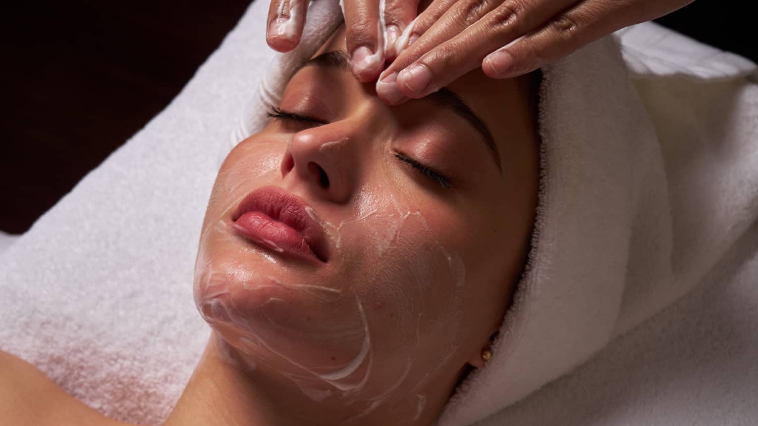 Person lies on massage table covered in a white cloth from shoulders down with hair wrapped in white towel while a pair of hands massages their forehead