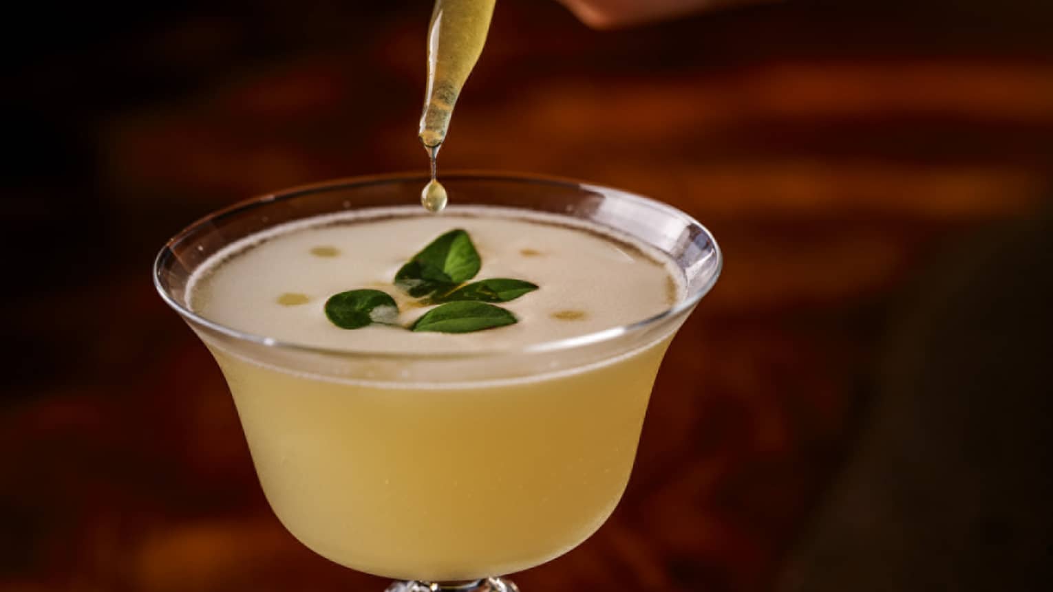 Eye dropper drips yellow liquid into frothy yellow cocktail with green herb garnish
