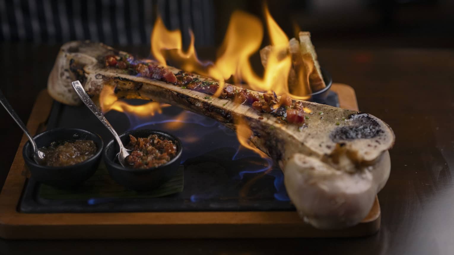 A large half of a bone is set aflame on a serving platter with two small, black bowls of toppings on the side