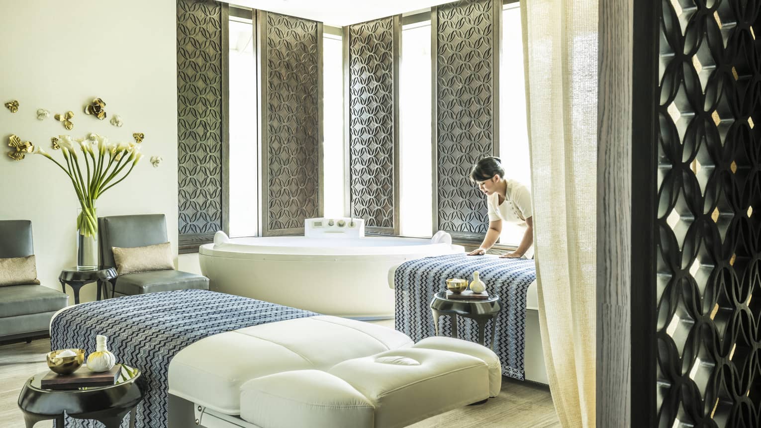Large white spa table in front of white spa tub, silver wall panels, woman places blanket on table