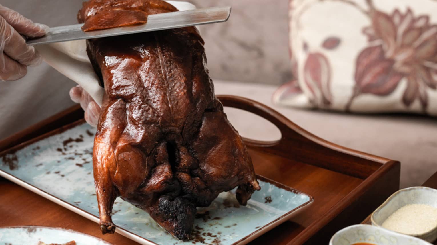 Chef's hands slicing a browned whole Peking duck over a rectangular dish on wood tray