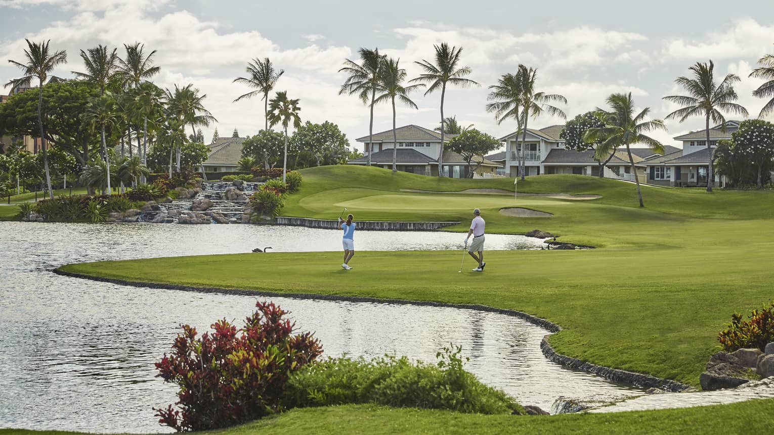 Two golfers on green grass near pond, palm trees in the distance