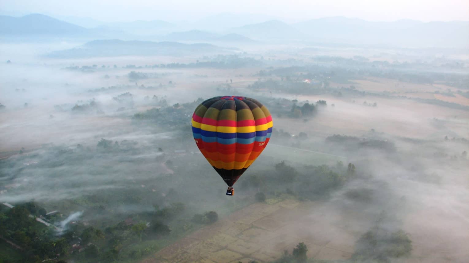 Colourful striped hot air balloon hovers high above field in clouds
