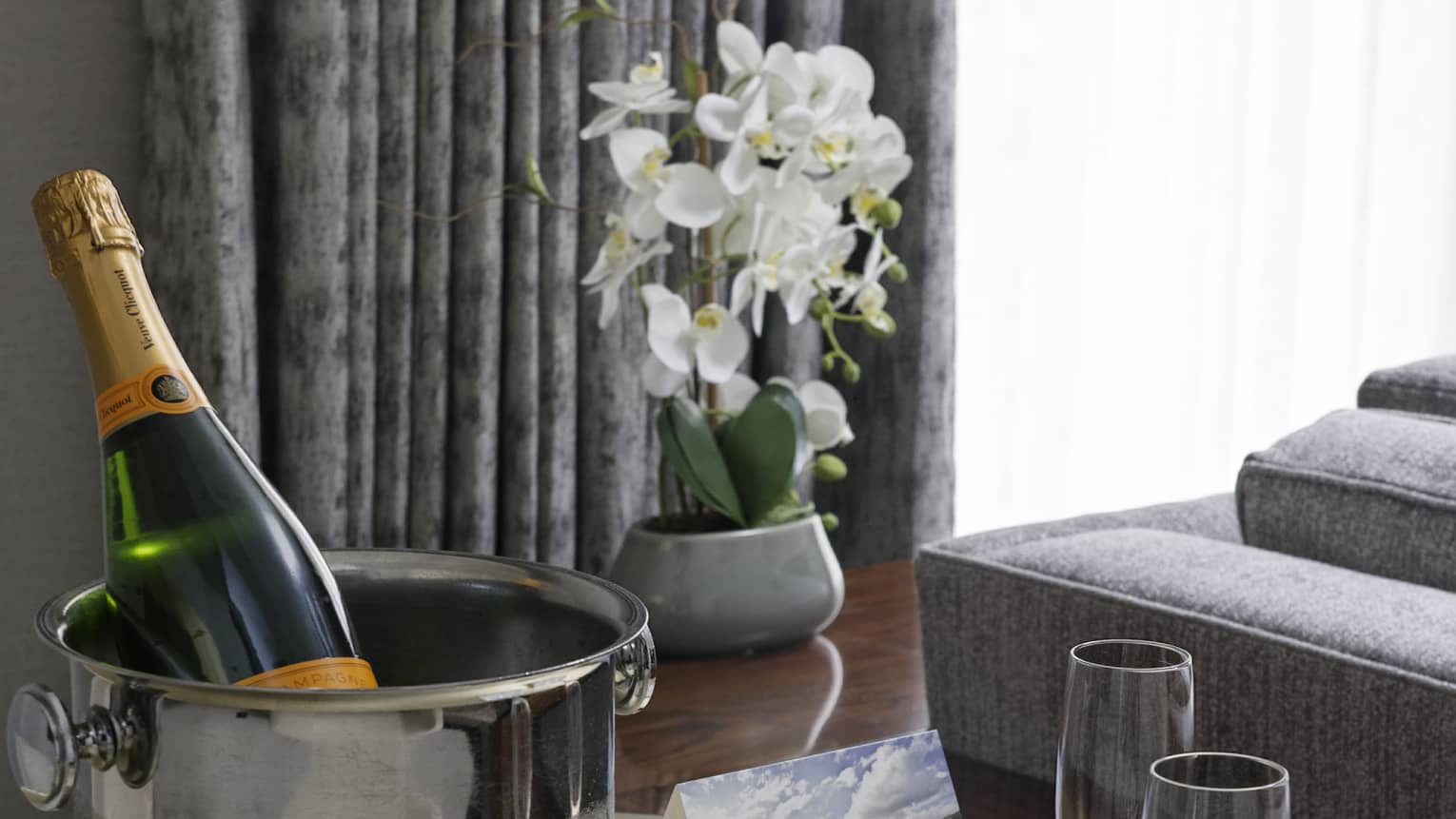 Ice bucket of Champagne and two flutes on round table, grey couch and window with grey curtains in the background