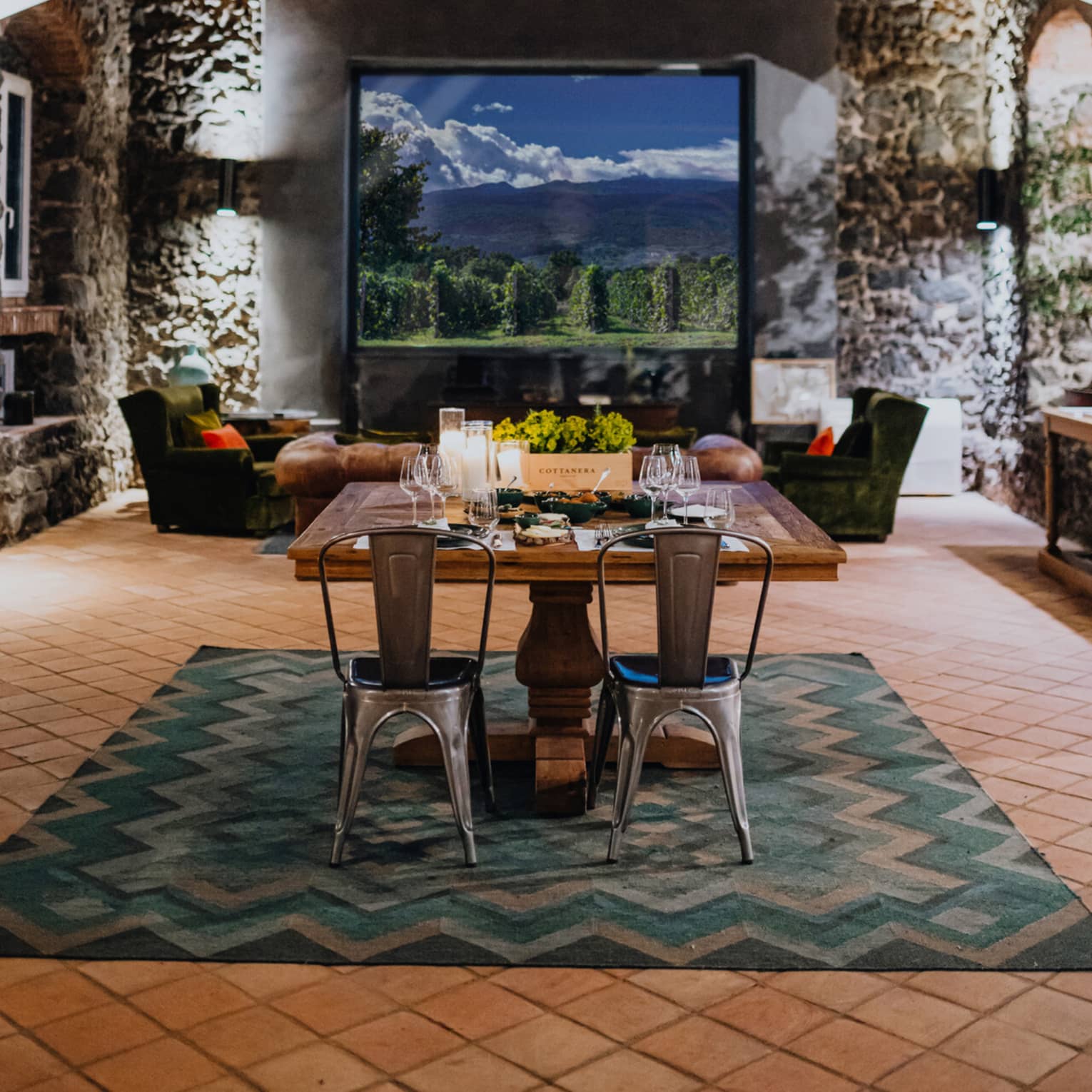 A wooden dining table set with floral arrangements and glassware, in a room with a window overlooking mountains and a vineyard