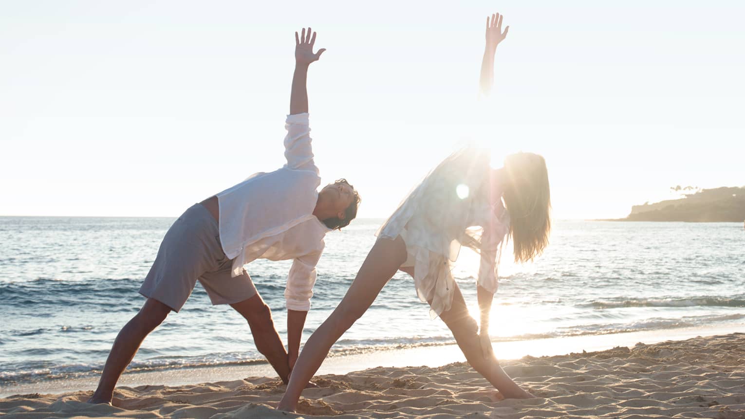 Two people in white beach shirts and shorts do a yoga pose with yogi at sunrise on beach