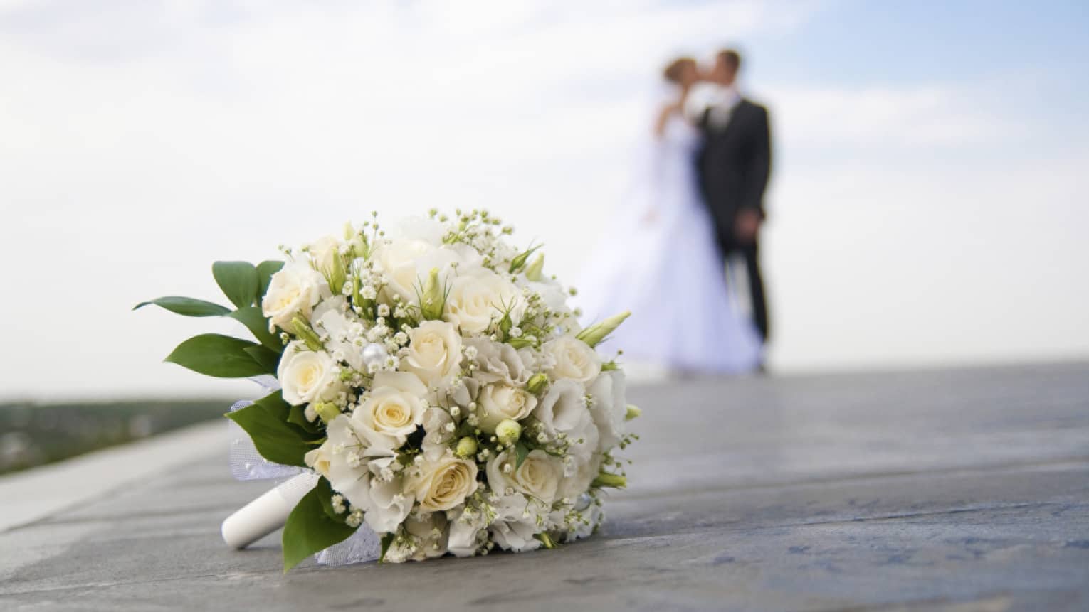 White floral wedding bouquet on ground, bride and groom kissing in background