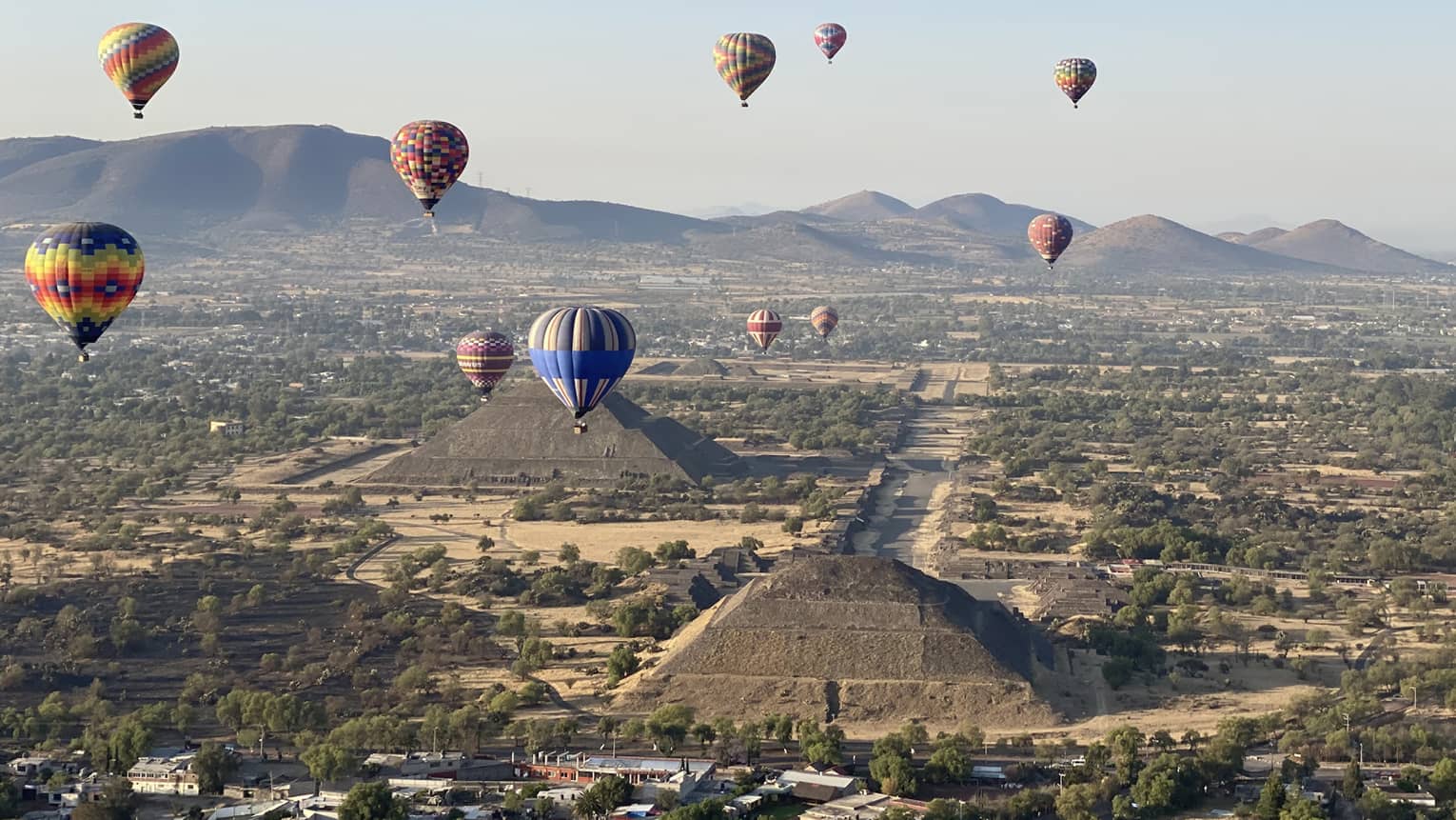 Hot-air balloons floating over old temples in a dessert covered in shrubs.