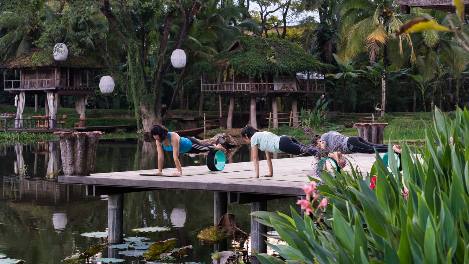 On a wooden deck above the water, a woman leads two people in a plank yoga move, with the feet balancing on a wheel
