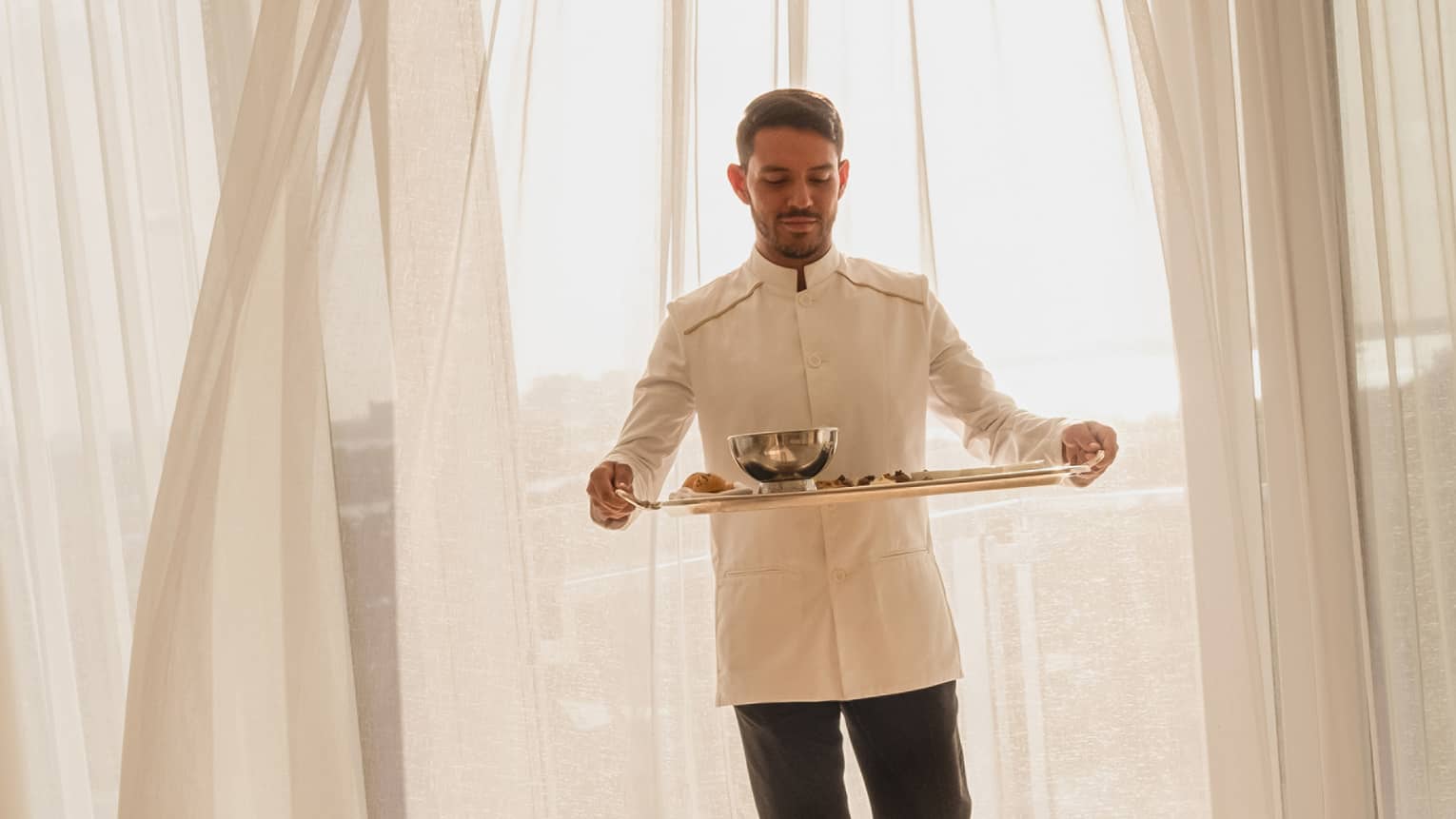 Hotel staff member carrying a tray with food for in-room dining, standing in front of sheer curtains with sunlight streaming through