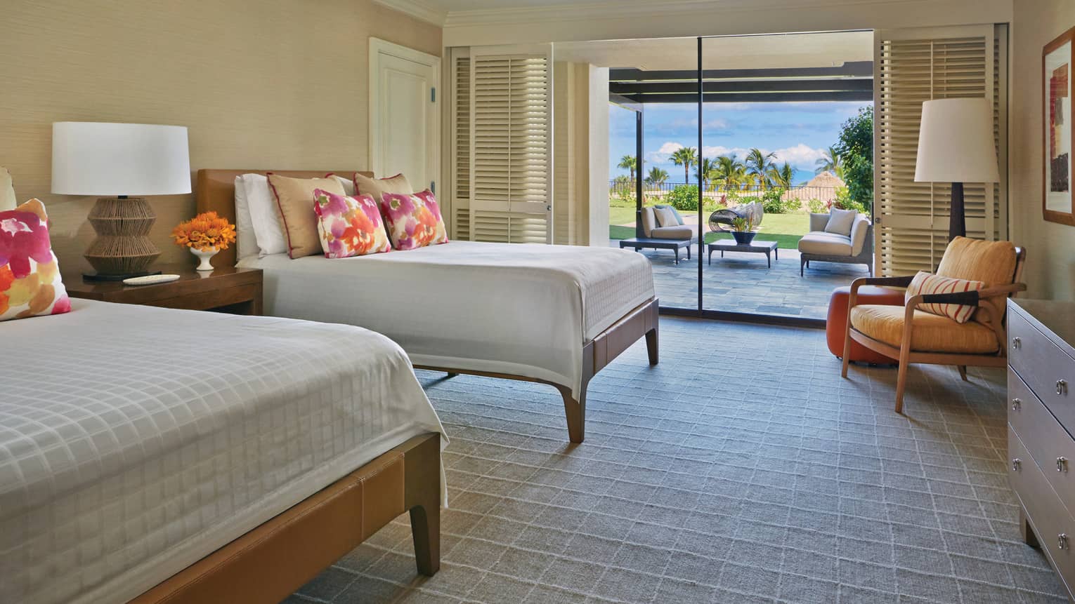 Bright guest room with two beds, colourful pillows, armchair and sliding doors opening to patio with garden view