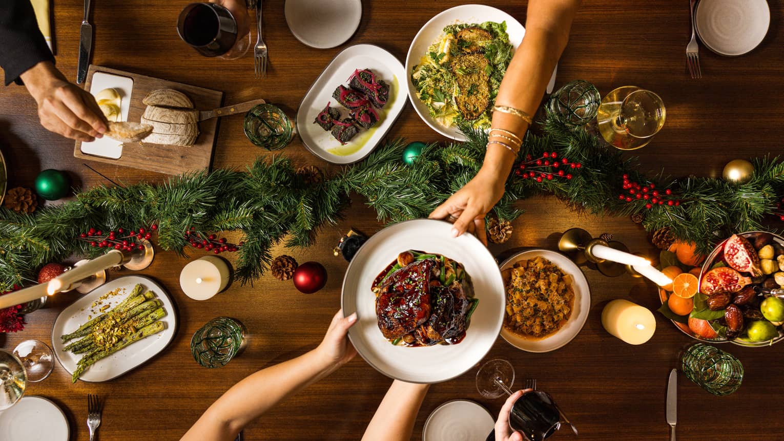 Hands pass a dish of food to another over a table set for a holiday meal with a green garland in the centre