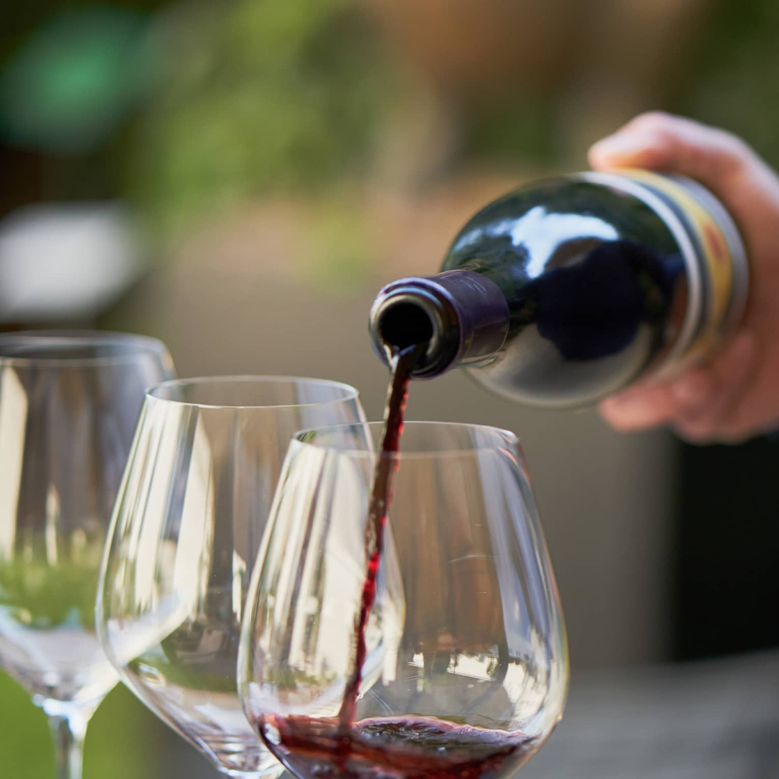 A close up of hands pouring a bottle of red wine into a clear wine glass that rests on a wooden tray, outdoors