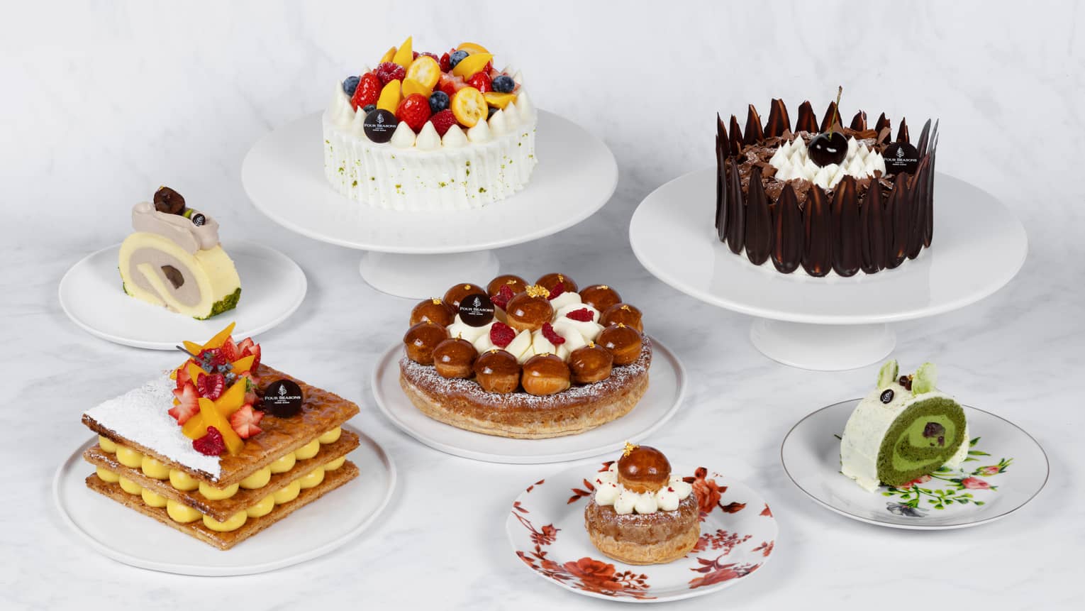 Assortment of gourmet desserts, including cakes topped with fruits and chocolate, cream-filled pastries and decorative layered treats on white plates.