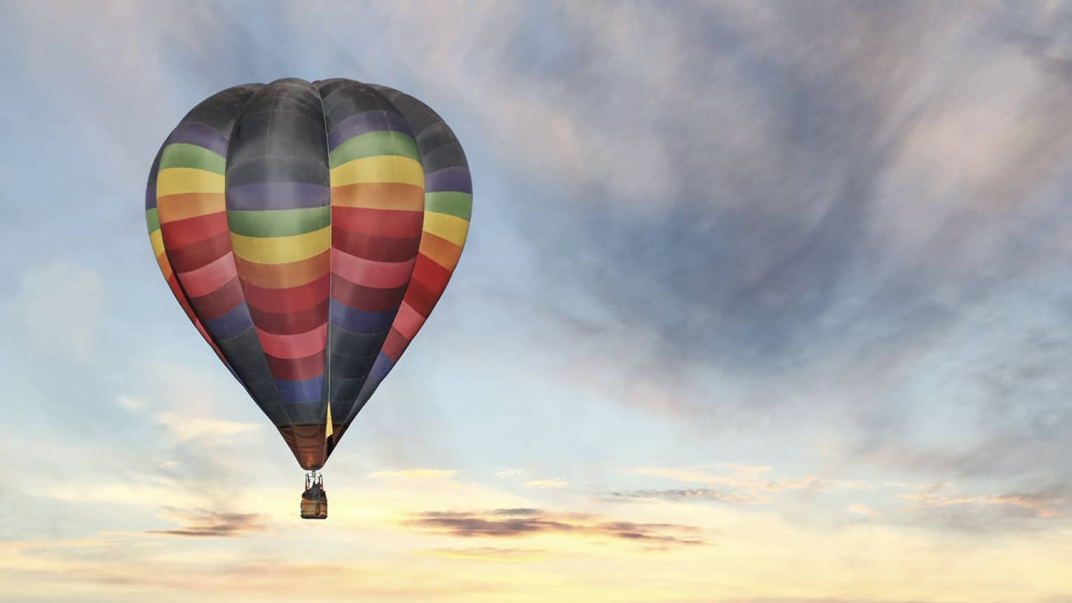Rainbow-striped hot air balloon floats in the sky at sunset