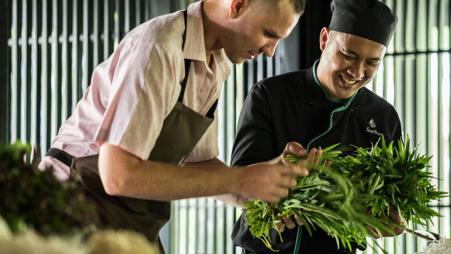 Smiling Chef Tran Van Sen holds greens, shows them to man in apron