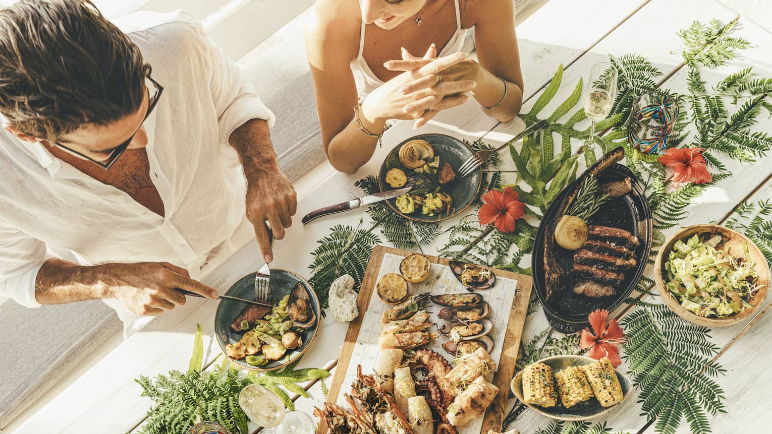A couple at a table of seafood canapés, grilled meat, corn and champagne flutes, adorned with palm fronds and red hibiscus.