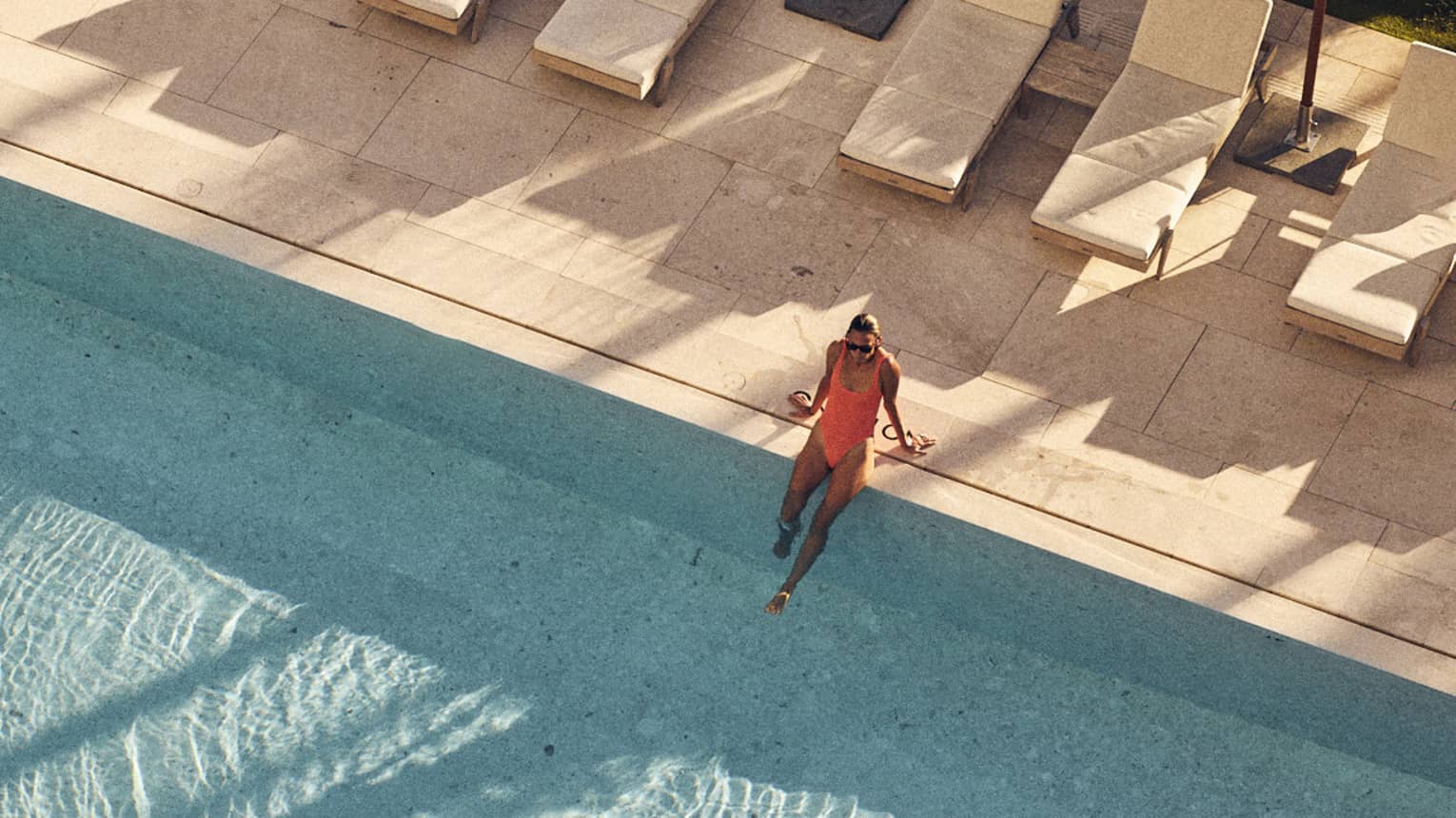 Aerial view of a woman in an orange swimsuit sitting on the edge of a large pool surrounded by lounge chairs and white umbrellas