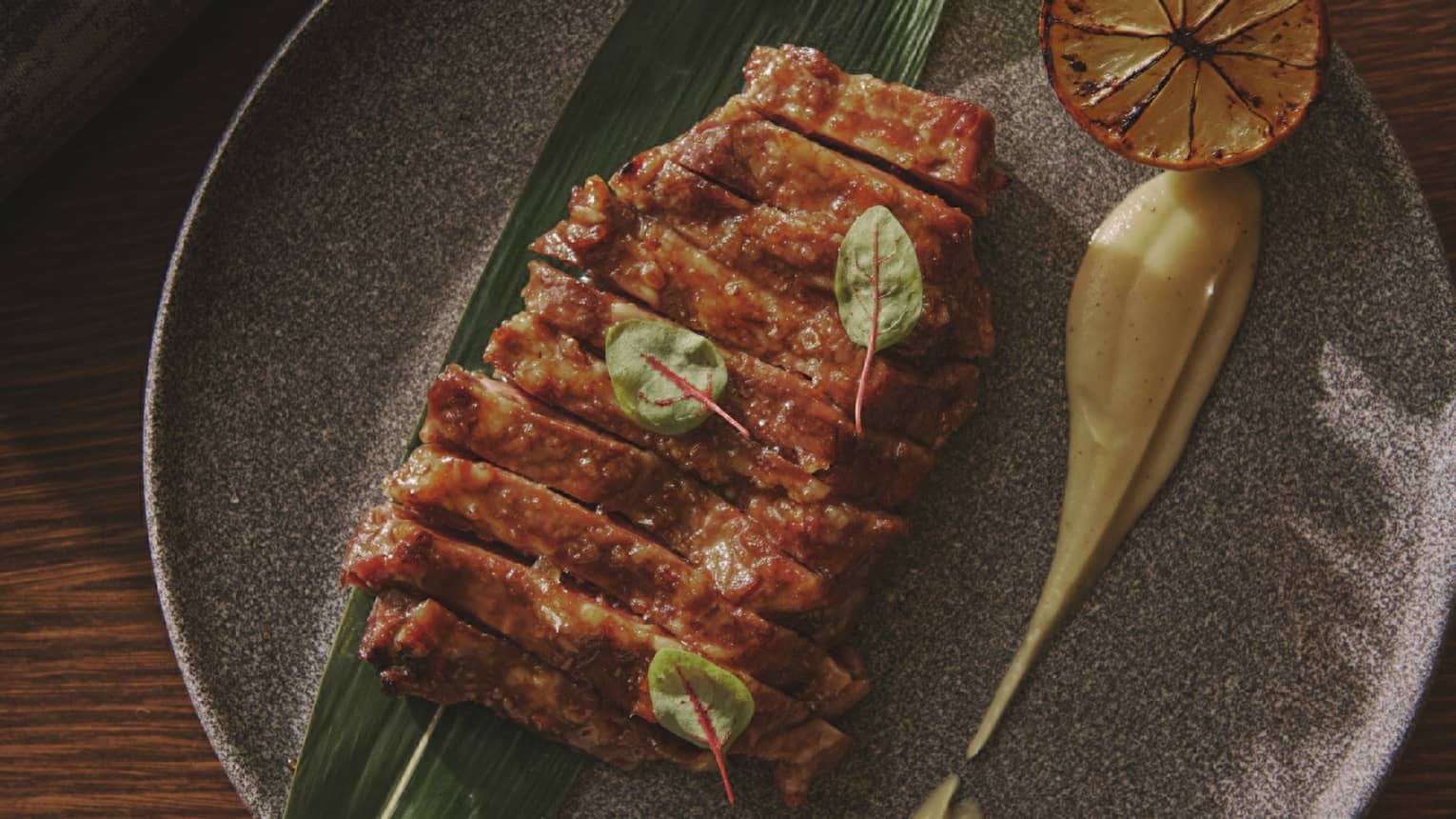 Sliced Pork with Greens Garnish atop long green leaf with Charred Lemon and ecru-coloured sauce on round ceramic plate