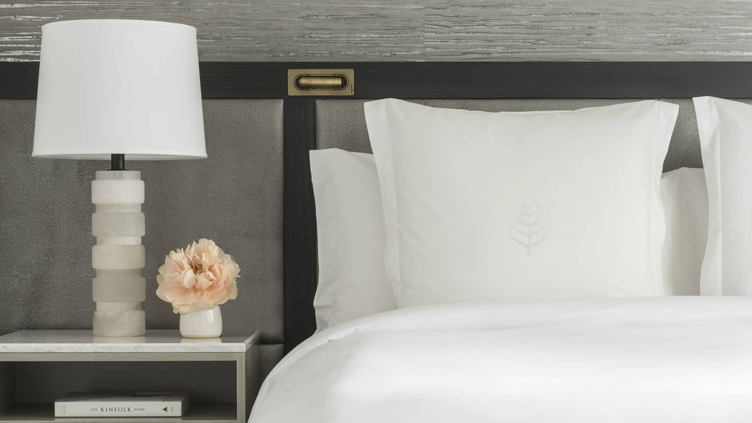 Luxury hotel room king bed with white sheets and Four Seasons tree logo embroidered on sham, next to a nightstand with white lamp and small vase of peach-coloured flowers